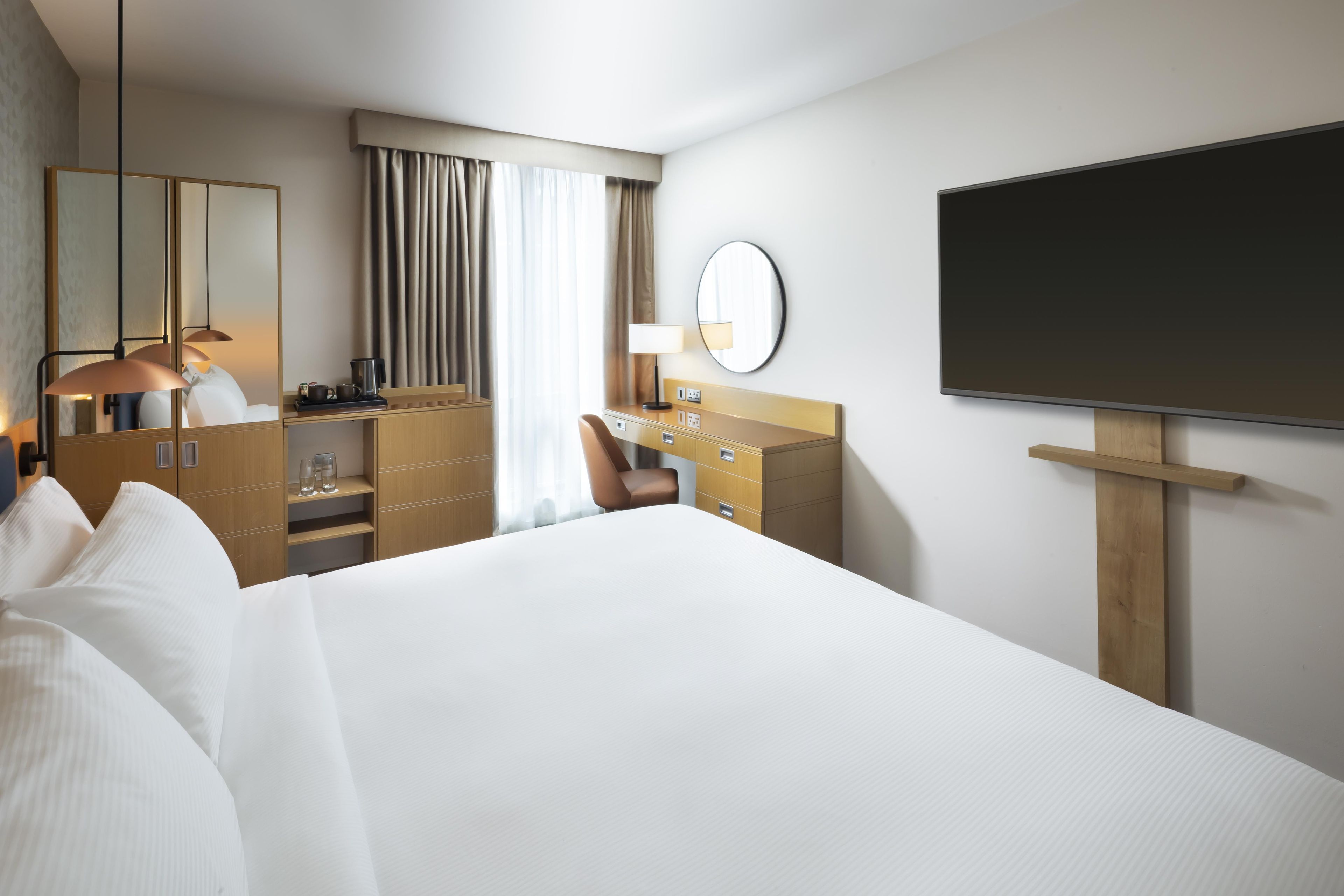 A hotel room with a large bed, a desk with a chair, a television, a mirror, and a window with curtains.