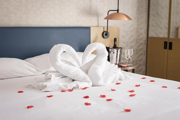 Cama bem arrumada com toalhas brancas dispostas em formato de coração, uma garrafa de champanhe e copos em uma mesa de cabeceira.