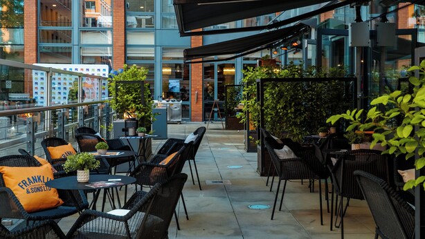 An outdoor café area with wicker chairs and tables, surrounded by greenery and modern buildings. - Hilton Garden Inn Birmingham Brindley Place (Birmingham)