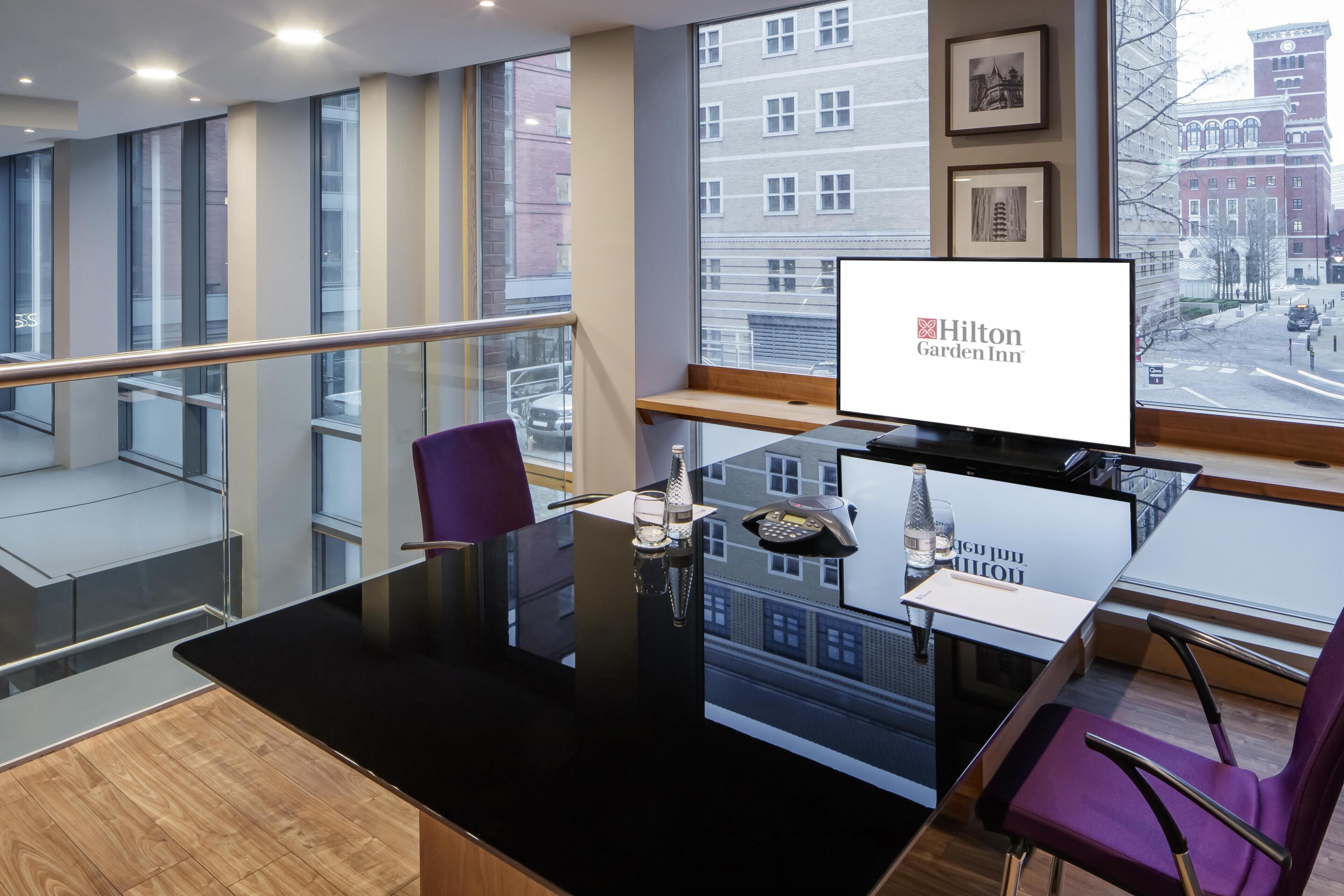 A modern hotel room with a glass table, purple chairs, a city view, and a large window.