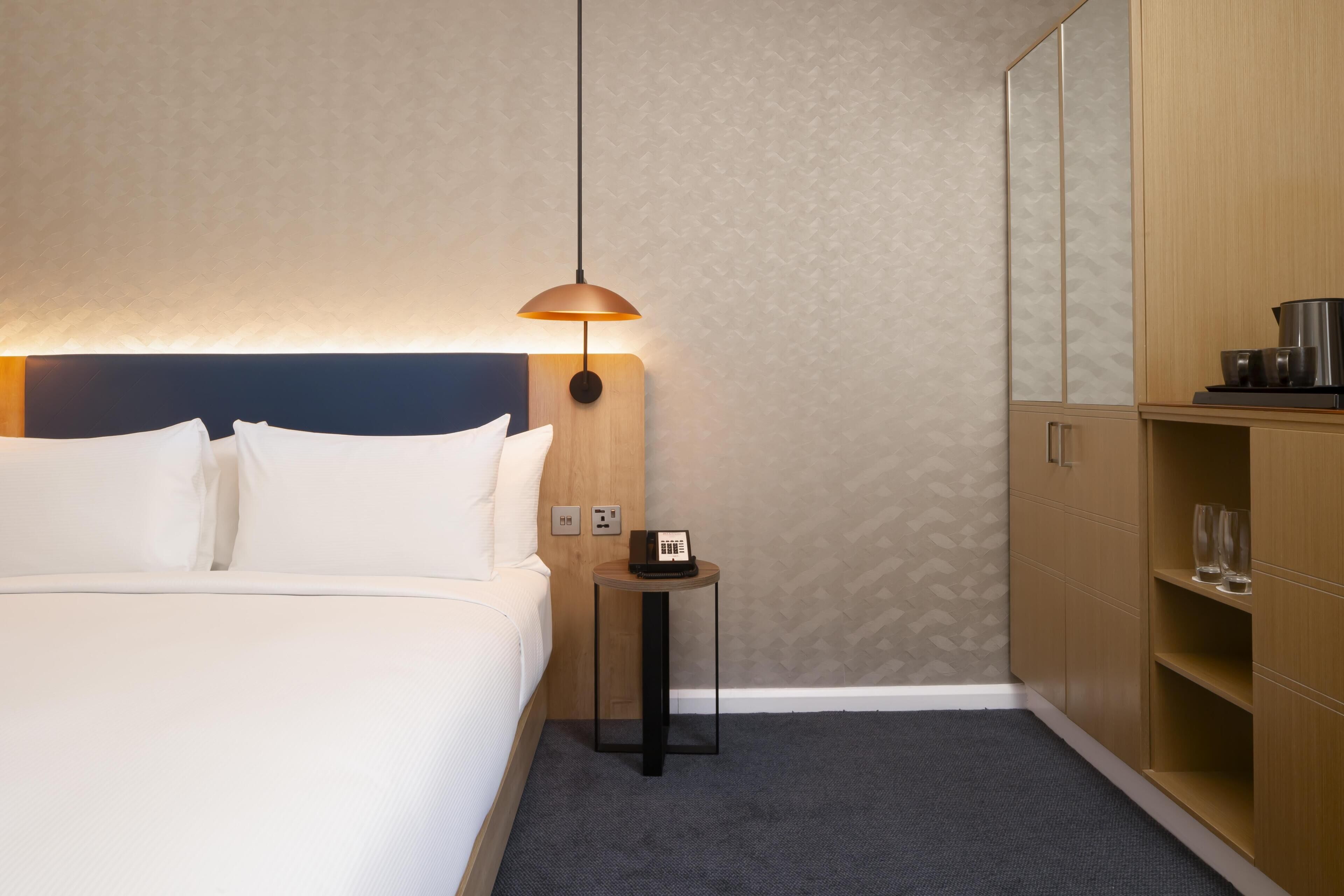 A hotel room with a bed, a bedside table with a lamp, a wooden wardrobe, and a small table with a kettle.