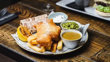 Une assiette de poisson-frites, accompagnée d’un quartier de citron, de sauce tartare et de purée de petits pois, posée sur une table en bois.