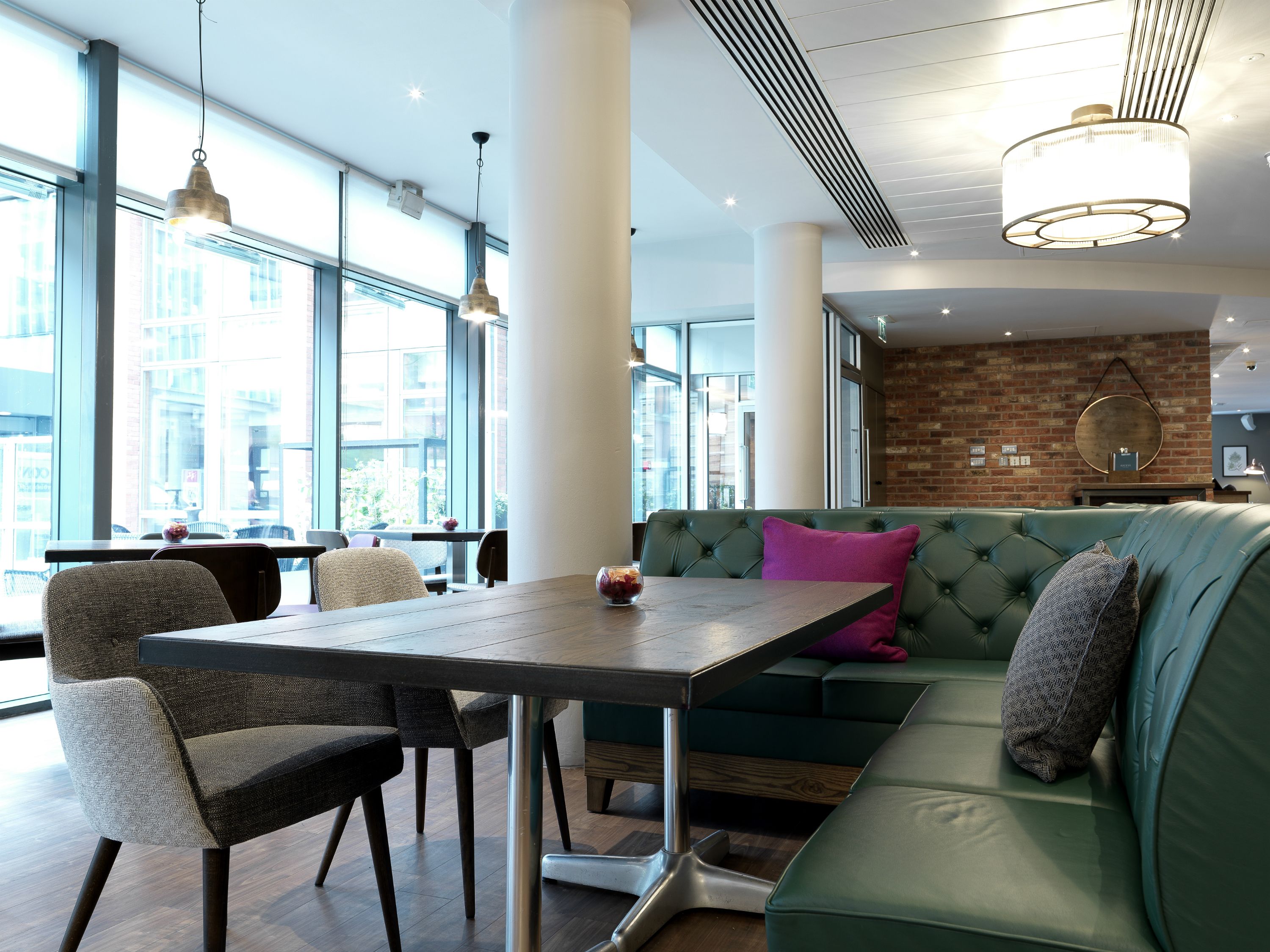 A modern interior with a green sofa, a dining table, and chairs. There are large windows providing natural light and a view of the city.