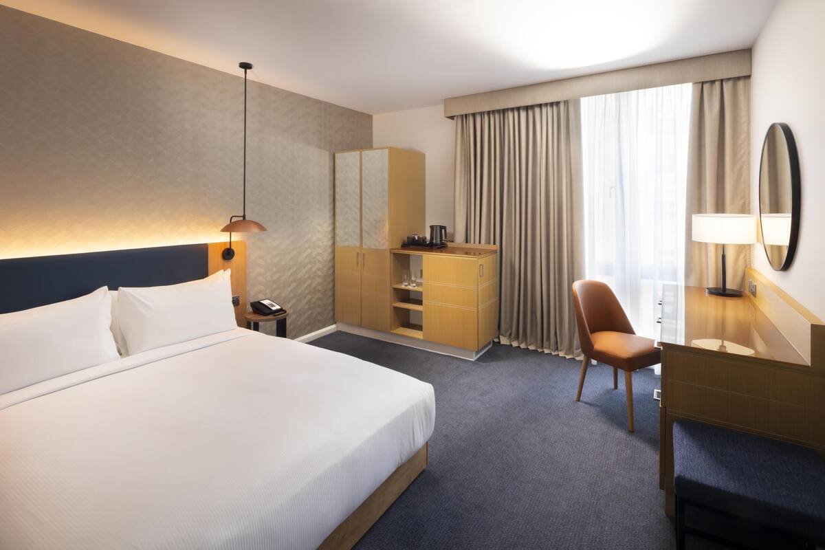 A hotel room with a large bed, a desk, a chair, a wardrobe, and a mirror.