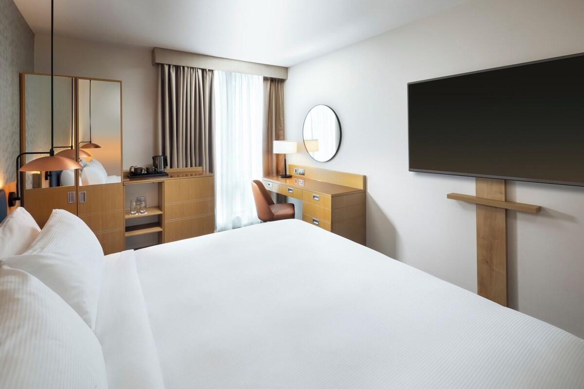 A hotel room with a large bed, a desk with a chair, a wardrobe, a mirror, a flat-screen TV, and a window with curtains.