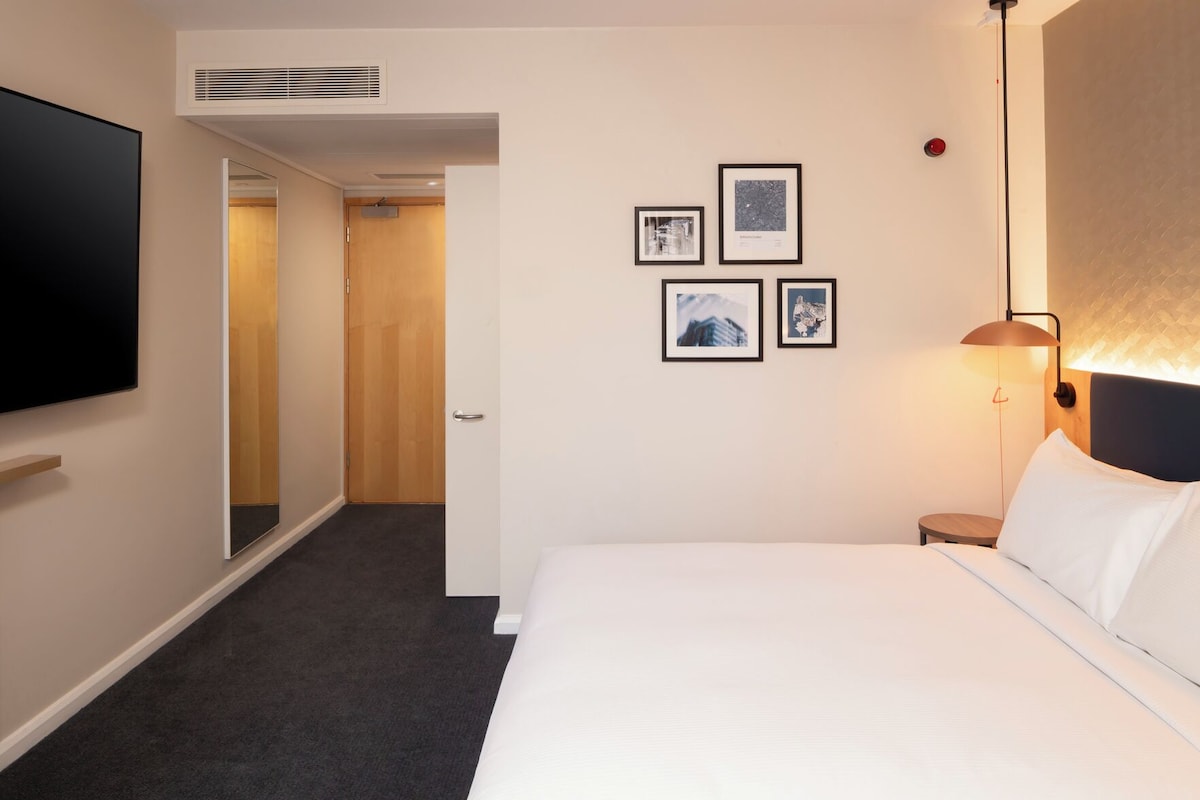 A hotel room with a bed, a flat-screen TV, a wall-mounted lamp, and framed pictures.