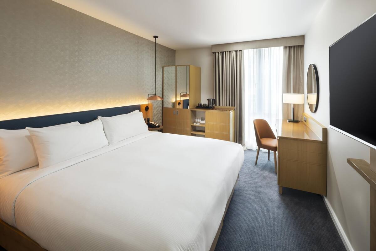 A hotel room with a large bed, a desk, a chair, a TV, and a window with curtains.