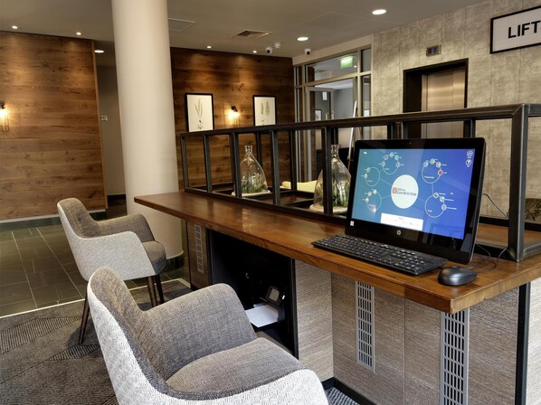 A hotel reception desk with a computer, two grey chairs, wooden panel walls, and a lift sign. - Hilton Garden Inn Birmingham Brindley Place (Birmingham)