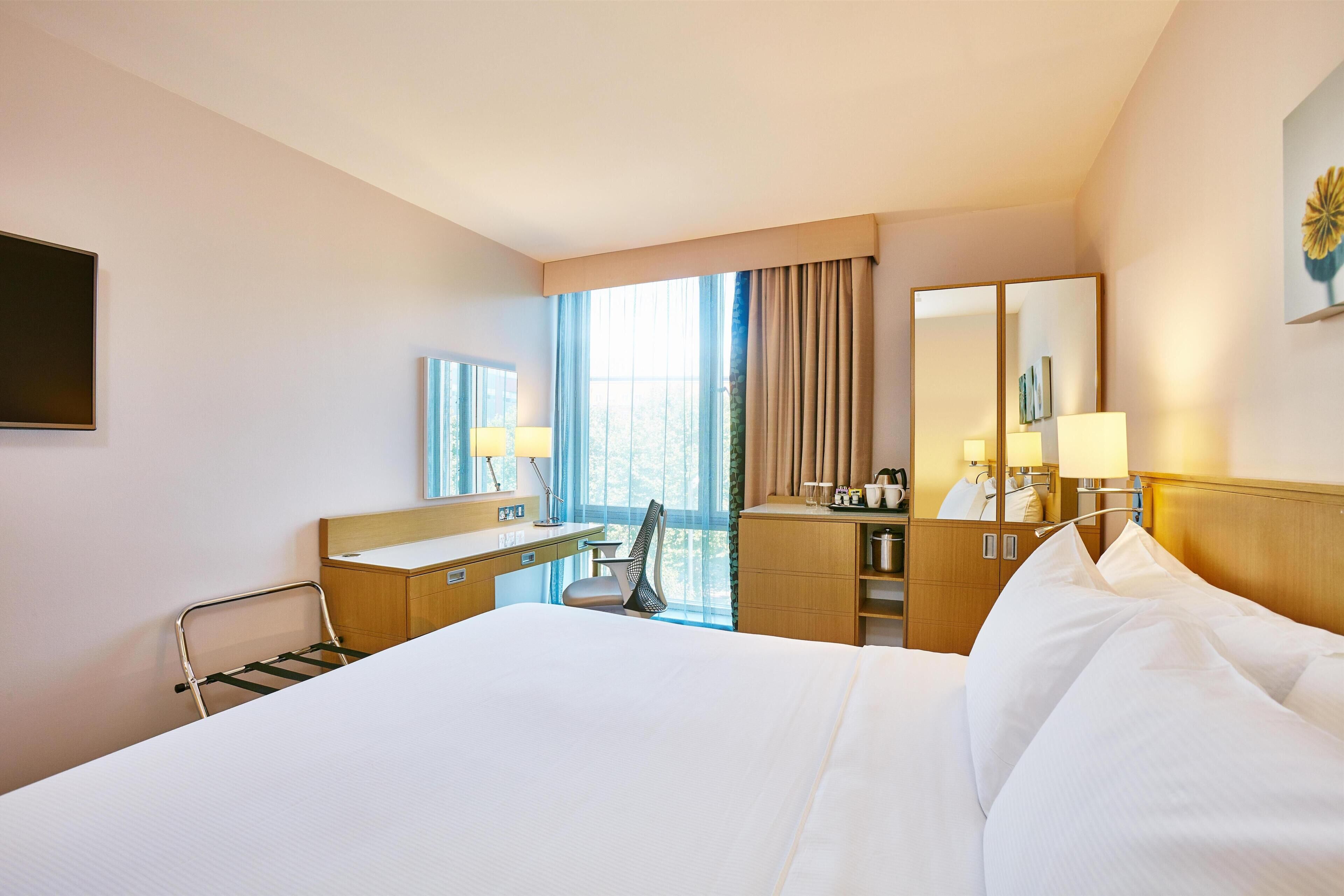 A hotel room with a bed, a desk, a chair, a wardrobe, a mirror, and a window with curtains.