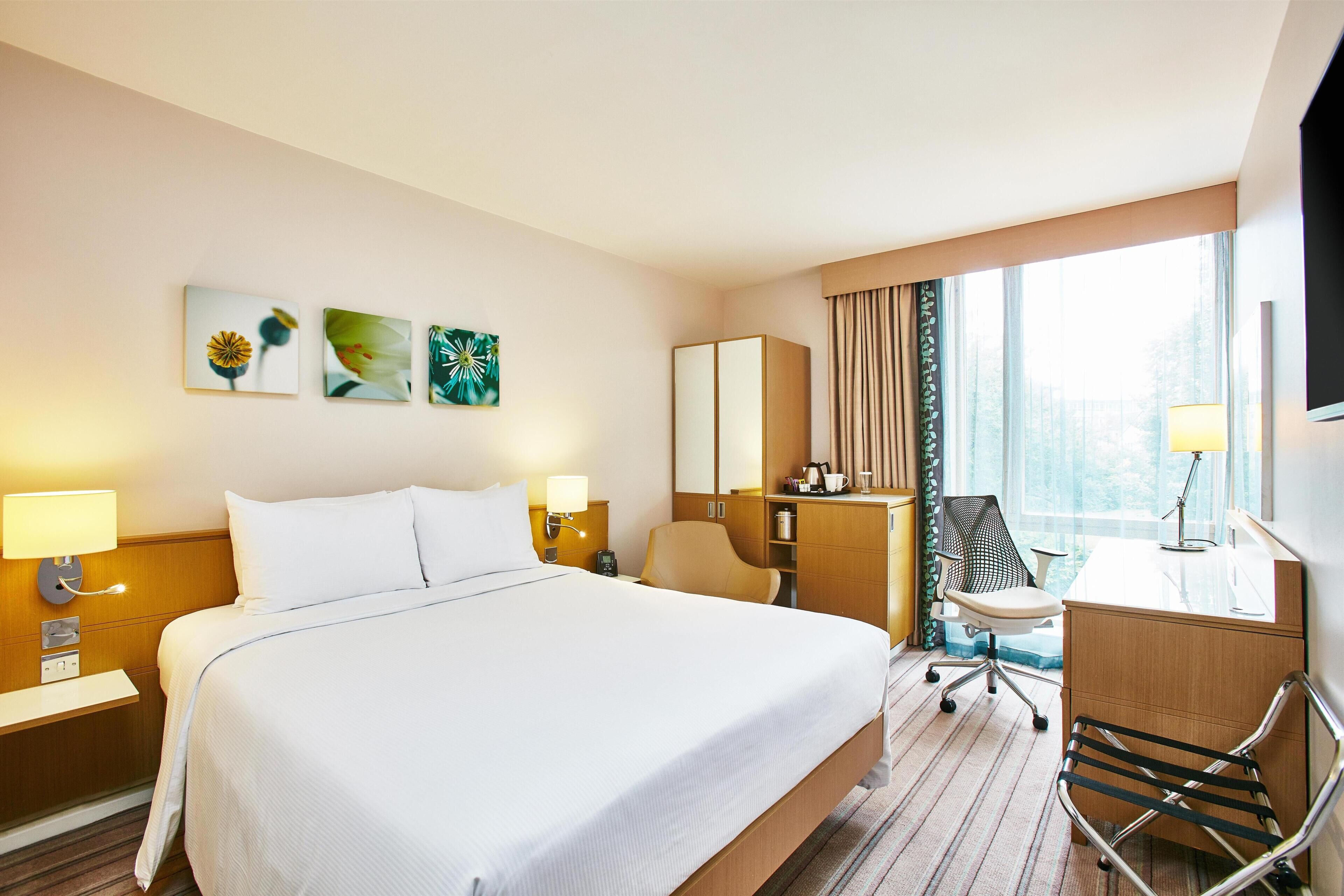 Habitación de hotel con una cama grande, un escritorio, una silla, una lámpara de escritorio, un televisor y obras de arte en la pared.