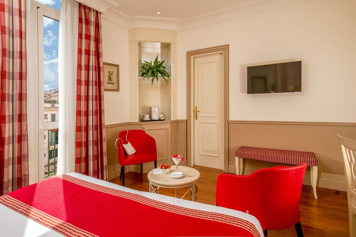 A hotel room with a red sofa, a small round table with a white tablecloth, a red armchair, a bench, a television mounted on the wall, and a window with red and white curtains.