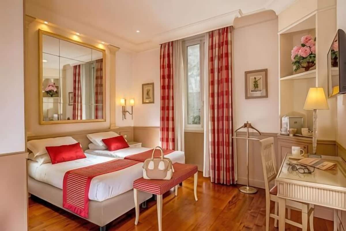 A hotel room with two beds, a desk, a chair, a mirror, a window with curtains, and a bedside table with a lamp.