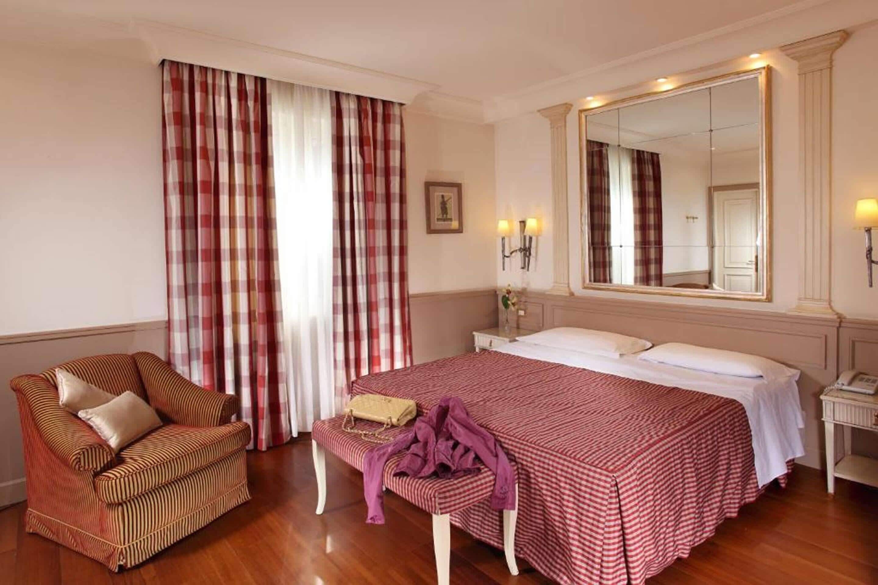 Une chambre d’hôtel avec un lit, une chaise, un banc, un miroir et des rideaux.