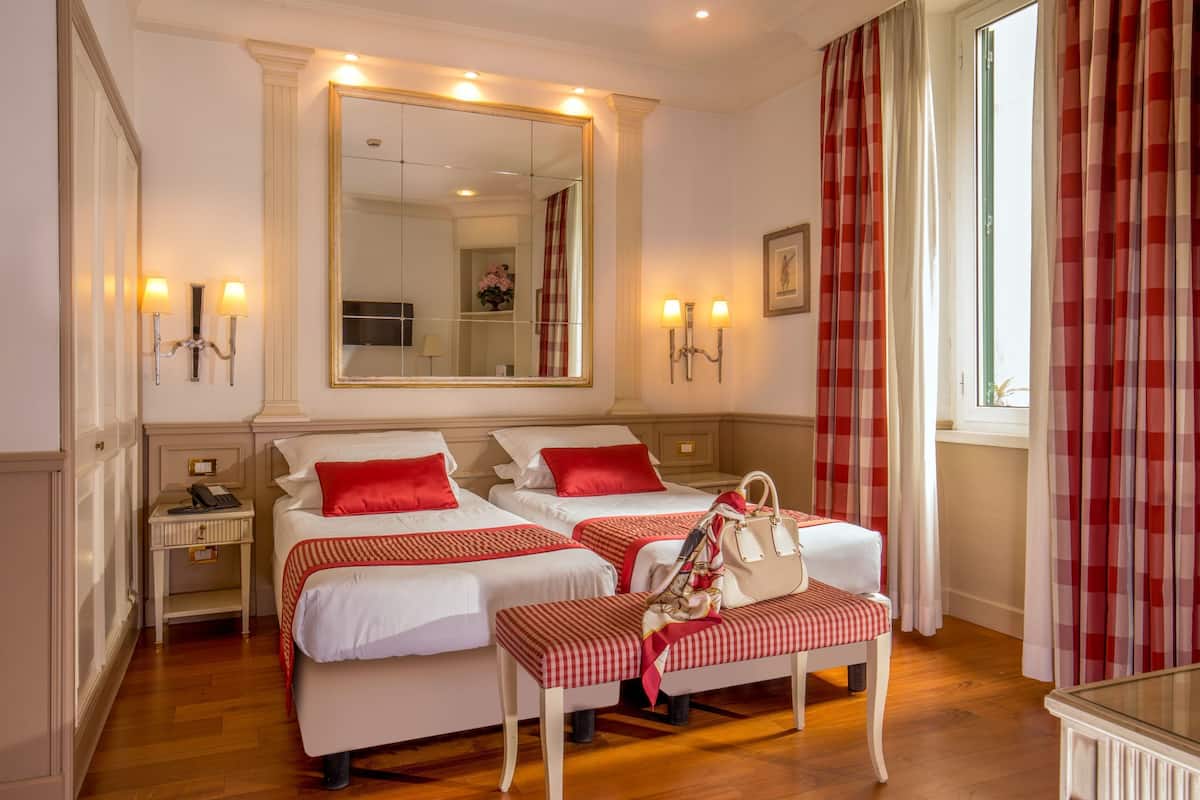 A hotel room with two beds, a bench, a mirror, and a window with curtains.