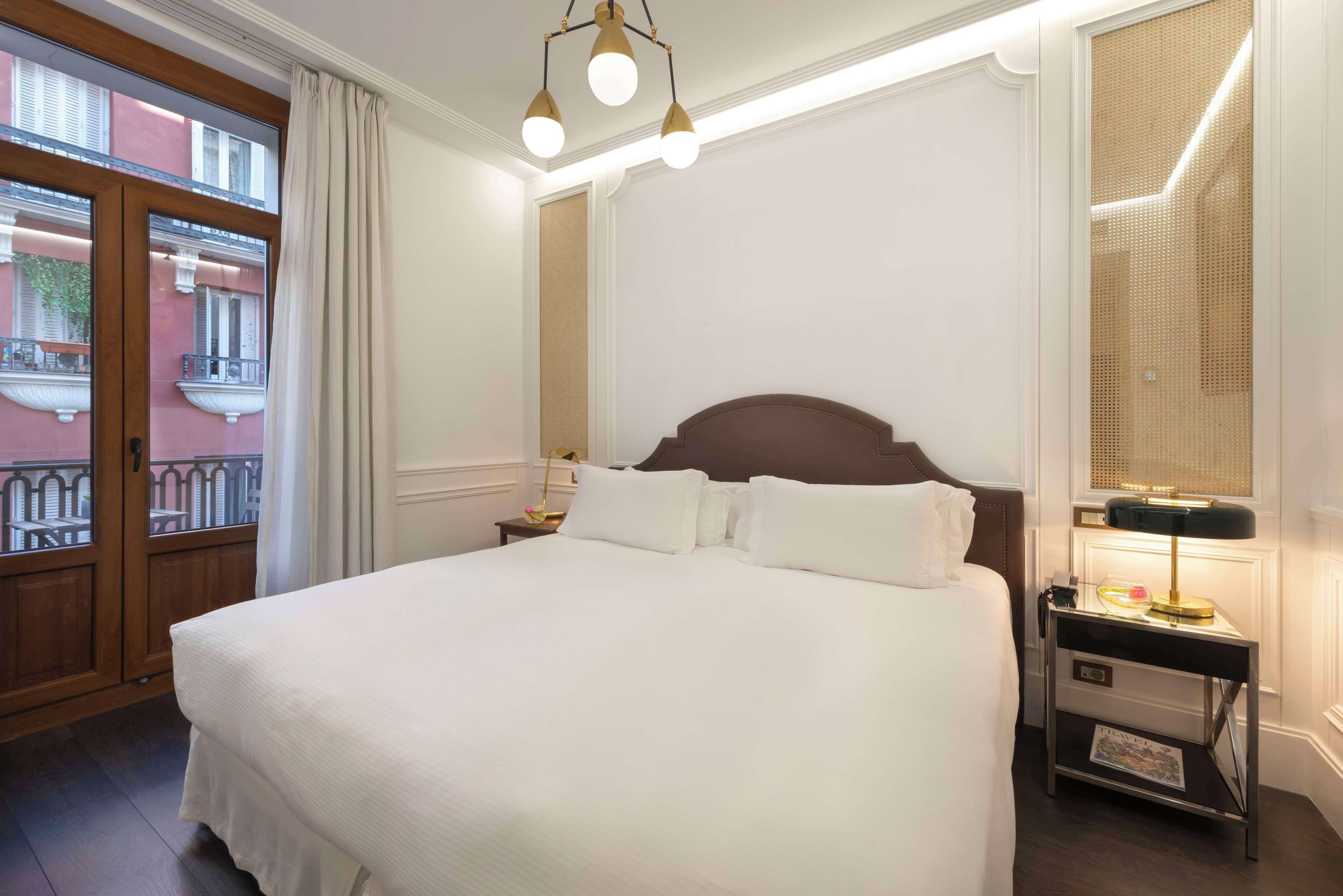 Un lit bien fait, avec du linge de lit blanc, une tête de lit en bois et une table de chevet avec une lampe. Une fenêtre avec des rideaux est visible, et un balcon avec une balustrade est aperçu à travers la fenêtre.