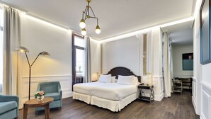 A hotel room with a large bed, two armchairs, a small table with a vase, a floor lamp, and a nightstand. - H10 Villa de la Reina Boutique Hotel (Madrid)
