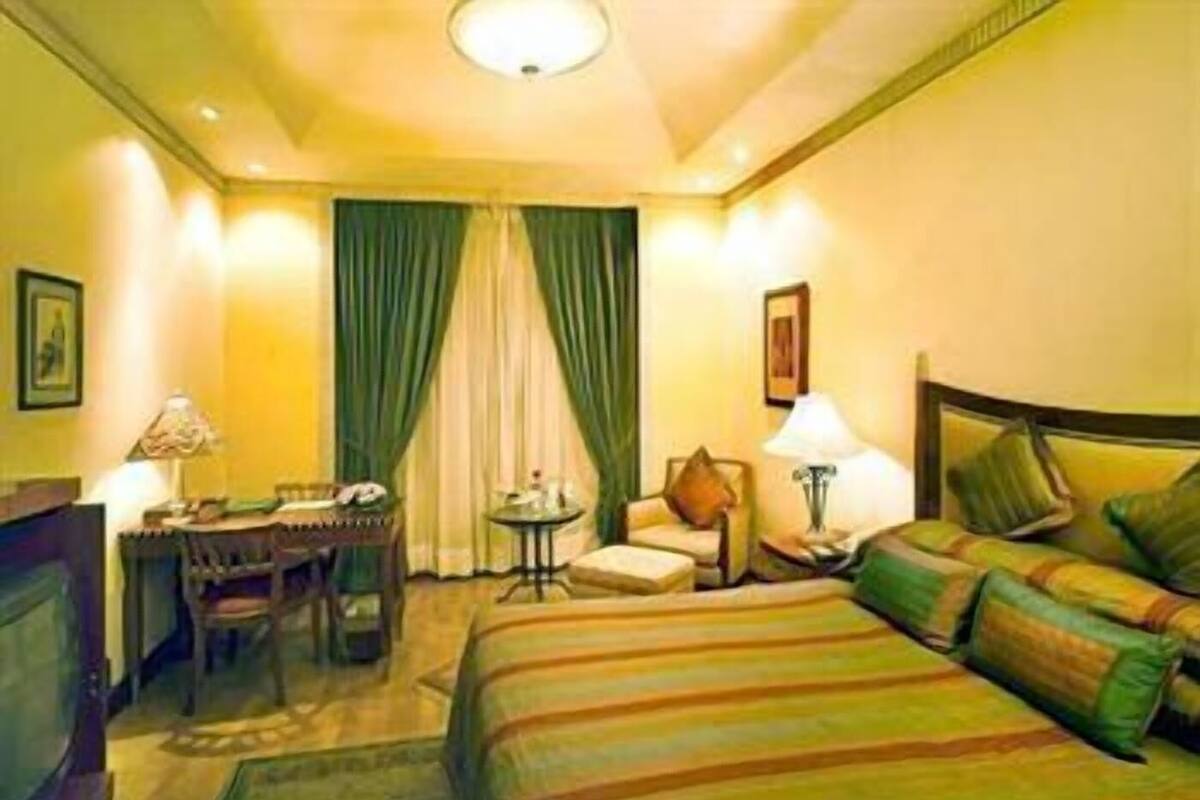 A hotel room with a bed, a desk, a chair, a small table, and a lamp.