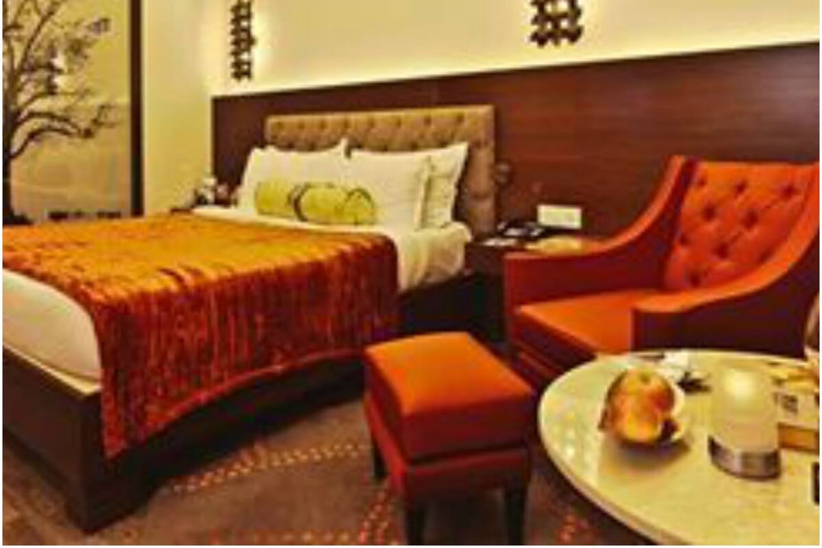 A hotel room with a large bed, a wooden headboard, a red armchair, a small table with a bowl of fruit, and a nightstand with a lamp.