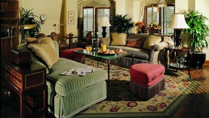 A spacious living room with a sofa, armchairs, a coffee table, and a dining table. There are potted plants, a clock on the wall, and a window with curtains. - The Rose Hotel (Pleasanton)