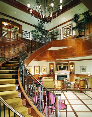 A grand staircase with ornate railings leading to an upper floor, a cozy sitting area with a fireplace, and a chandelier. - The Rose Hotel (Pleasanton)