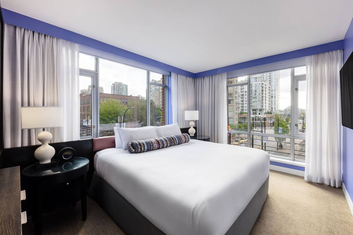 Una habitación de hotel moderna con una cama grande, dos mesitas de noche y vistas a la ciudad a través de amplias ventanas.