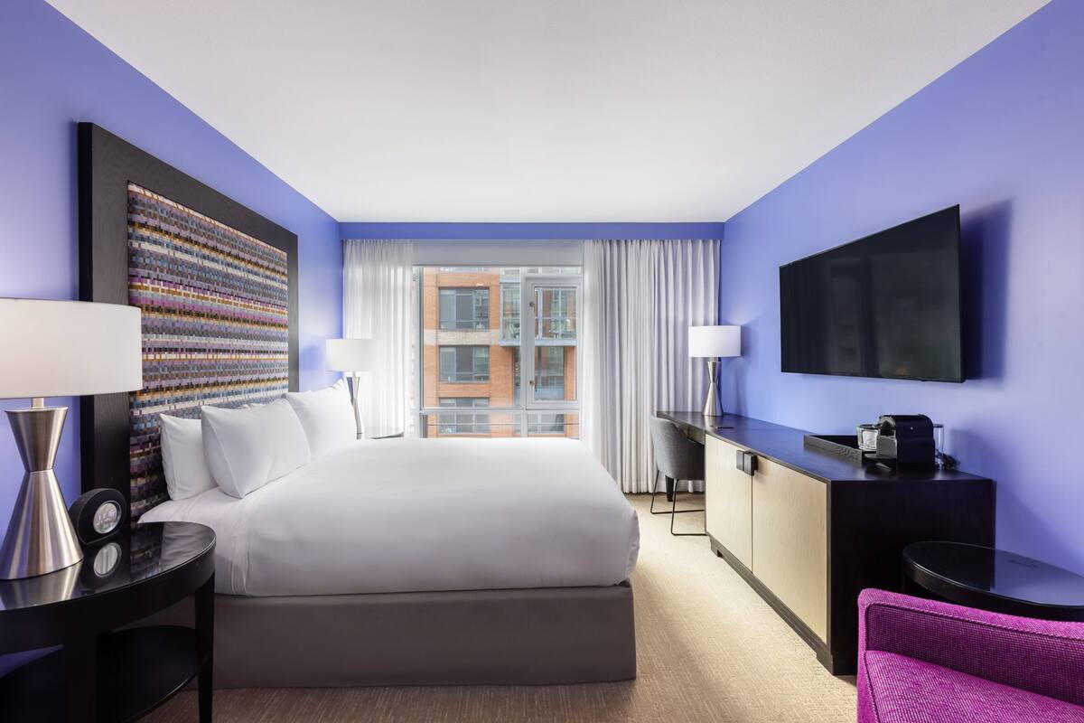 Habitación de hotel con una cama grande, un escritorio, una silla, televisión y vistas a edificios a través del ventanal.