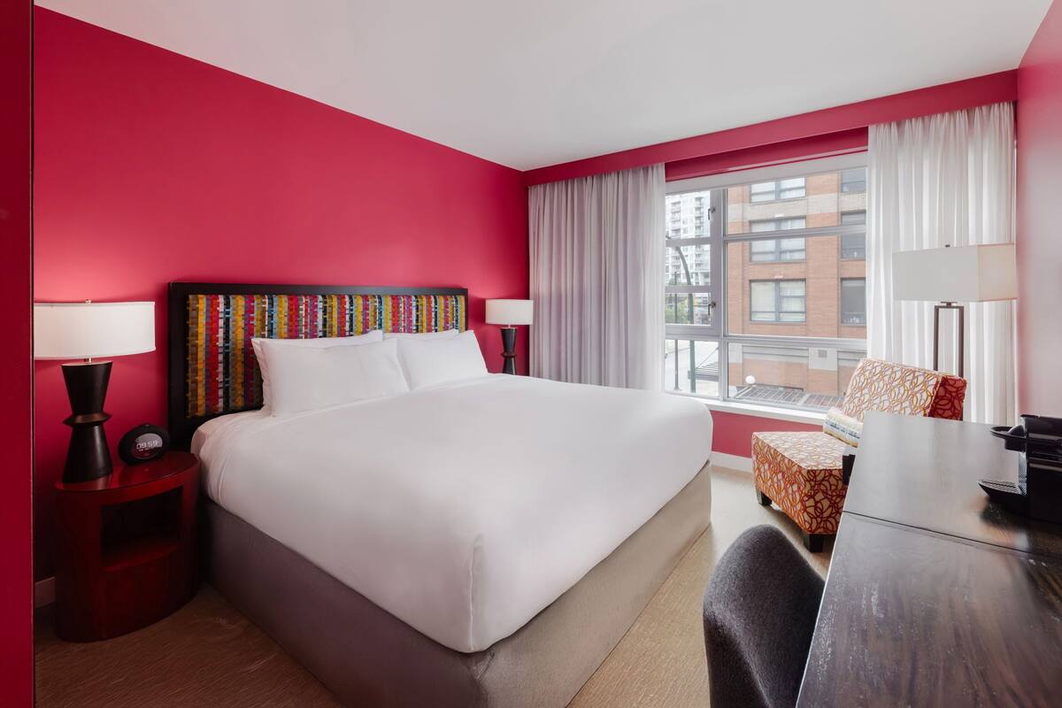 Habitación de hotel con una cama grande, un escritorio, una silla, una mesita de noche con lámpara y una ventana con cortinas.