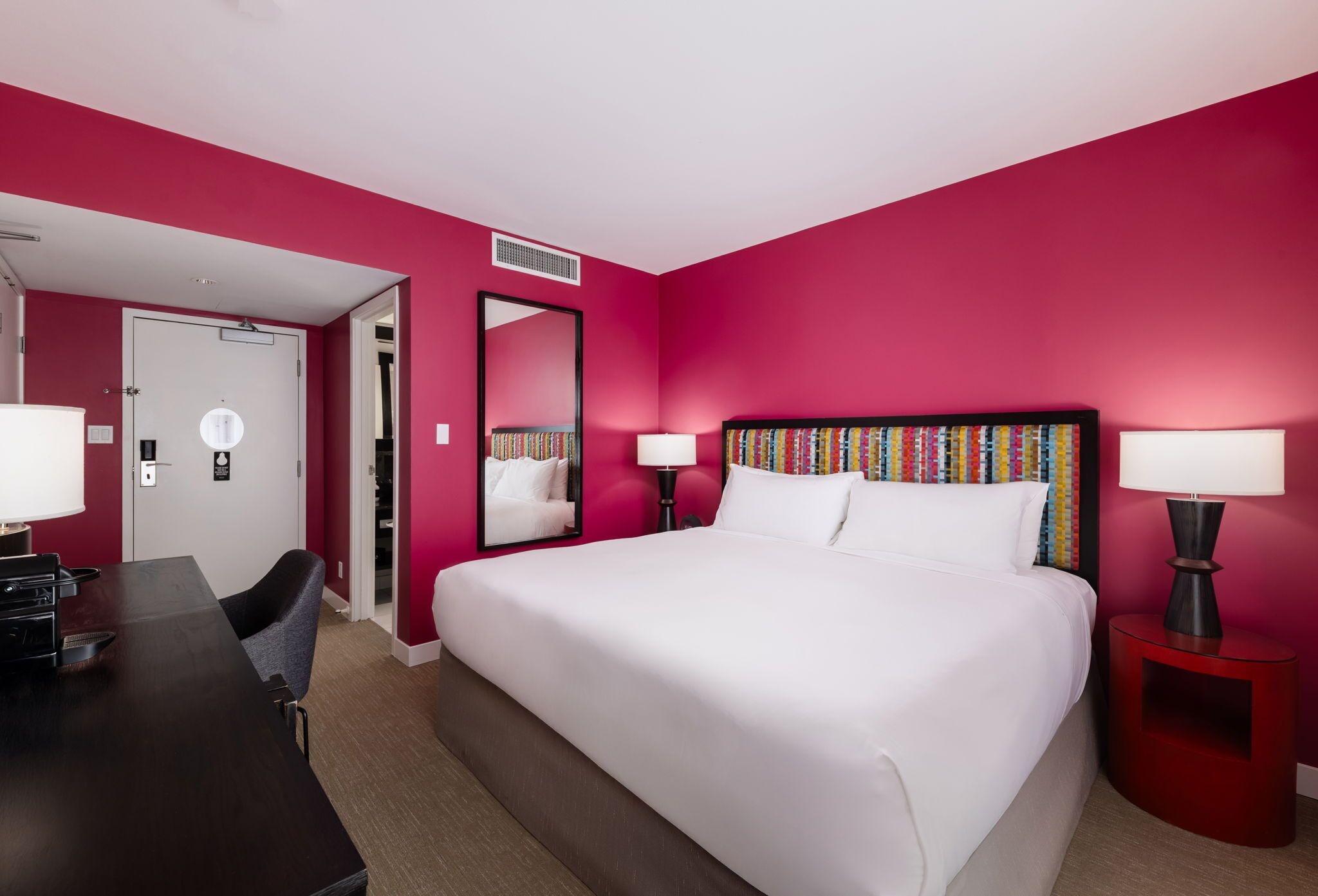 Ein Hotelzimmer mit einem großen Bett, einem Schreibtisch und einem Sessel. An der Wand befindet sich ein Spiegel und eine Bücherwand mit farbenfrohen Büchern.