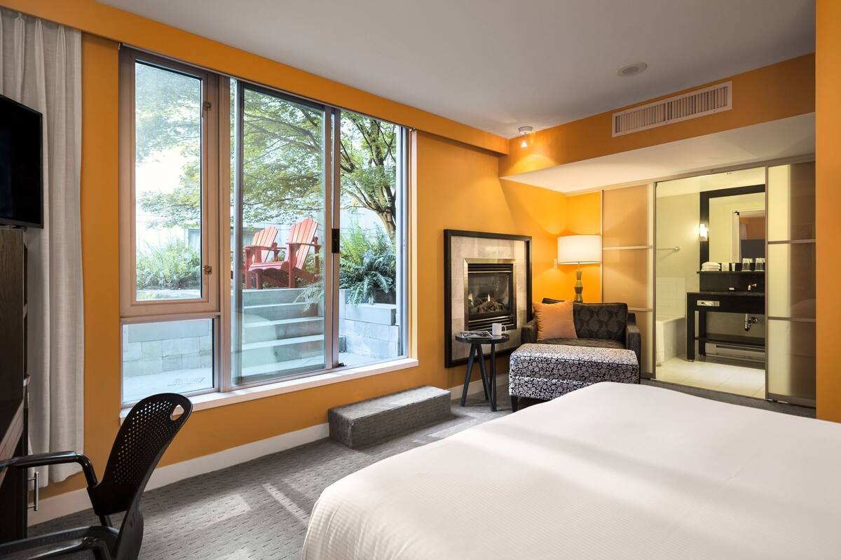 Habitación de hotel con cama, escritorio, silla, chimenea y un ventanal con vista a muebles de exterior.