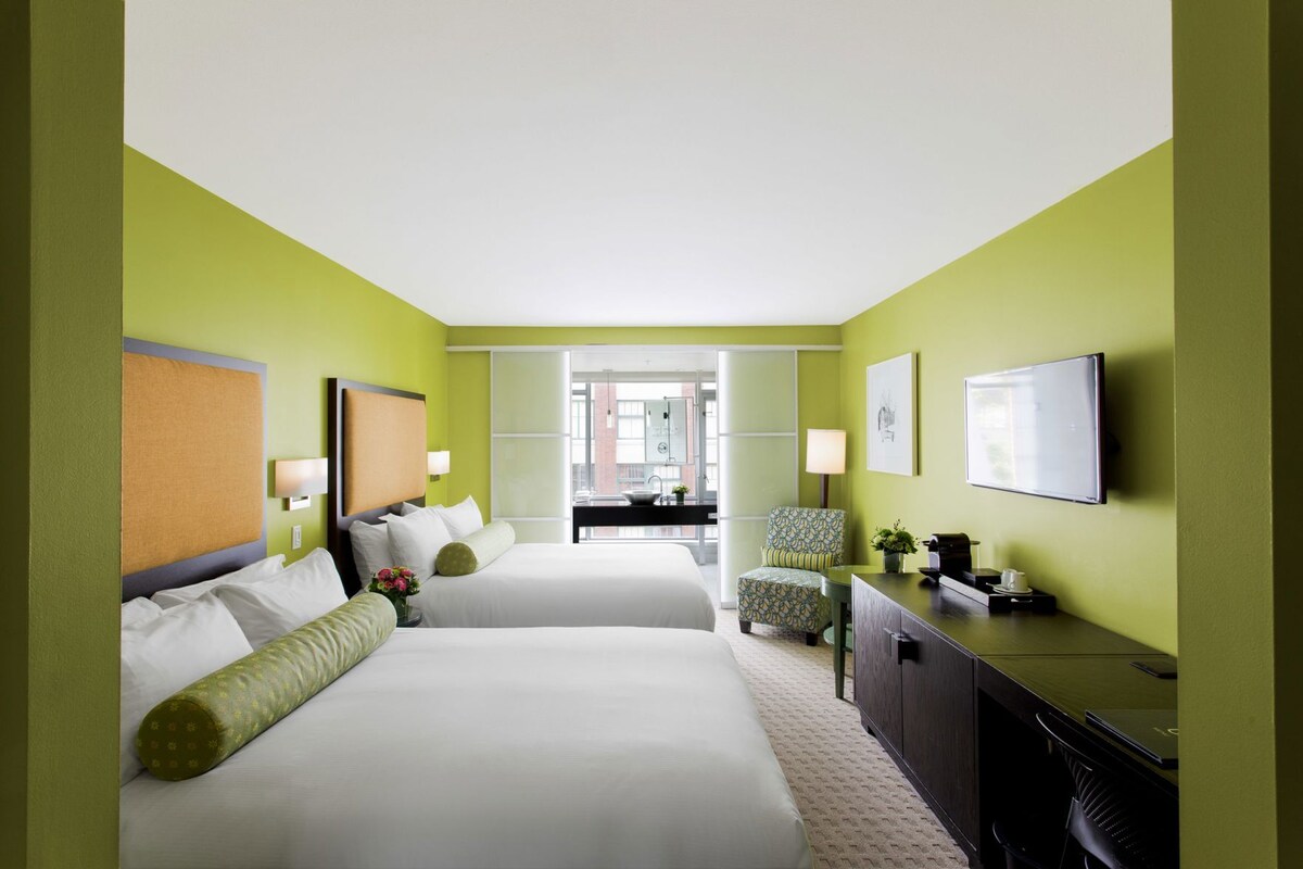A hotel room with two beds, a desk, a chair, a TV, and a view of the street.