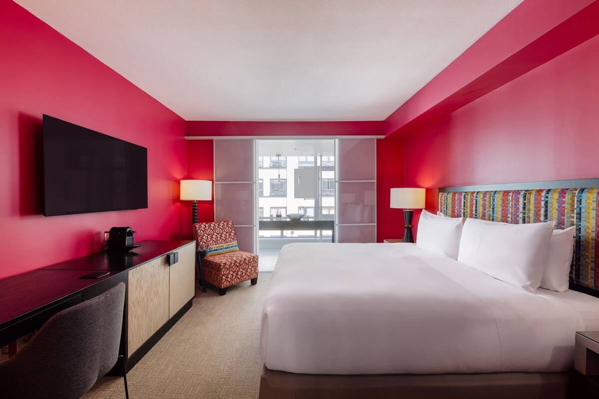 Habitación de hotel con una cama grande, un escritorio, un televisor de pantalla plana, una silla y una ventana con vista a edificios.