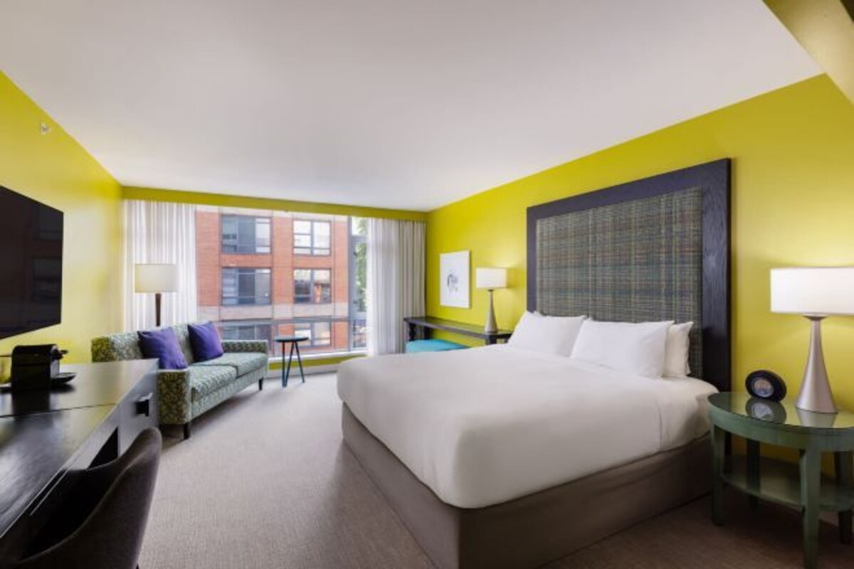 A hotel room with a bed, a desk, a chair, a sofa, a TV, a window with curtains, and a view of buildings.