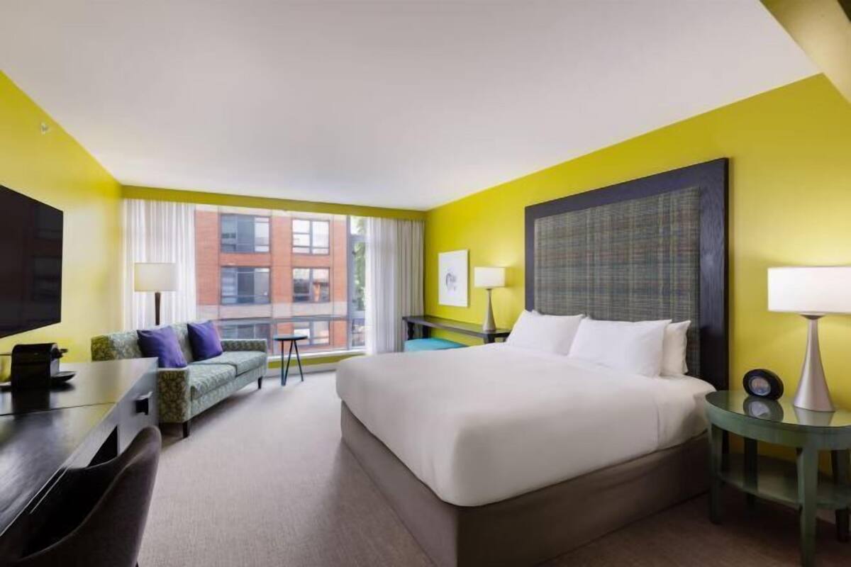 Habitación de hotel con cama, escritorio, silla, sofá, televisión y ventana con cortinas, con vista a edificios.