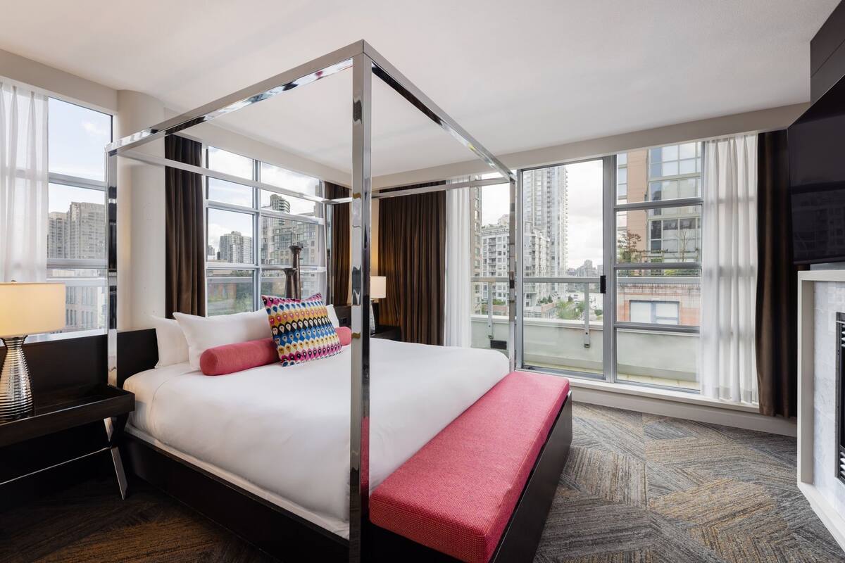 Una habitación de hotel moderna con una cama con dosel, ventanas amplias que ofrecen vistas de la ciudad, un escritorio con lámpara y un banco rojo.