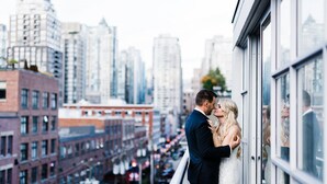 A couple kissing on a balcony overlooking a cityscape with tall buildings and a busy street. - OPUS Hotel Vancouver (Vancouver)