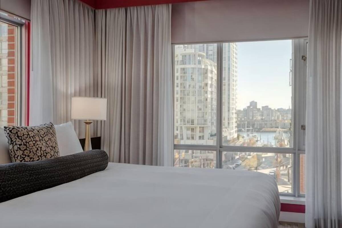 Habitación de hotel con una cama grande, una mesita de noche, una lámpara y vista a un paisaje urbano a través de un ventanal amplio.