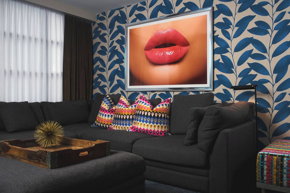 Una sala moderna con un sofá gris oscuro, cojines estampados y coloridos, una bandeja de madera con un objeto decorativo y un cuadro grande enmarcado con la imagen de labios en la pared.
