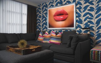 Una sala moderna con un sofá gris oscuro, cojines estampados y coloridos, una bandeja de madera con un objeto decorativo y un cuadro grande enmarcado con la imagen de labios en la pared.