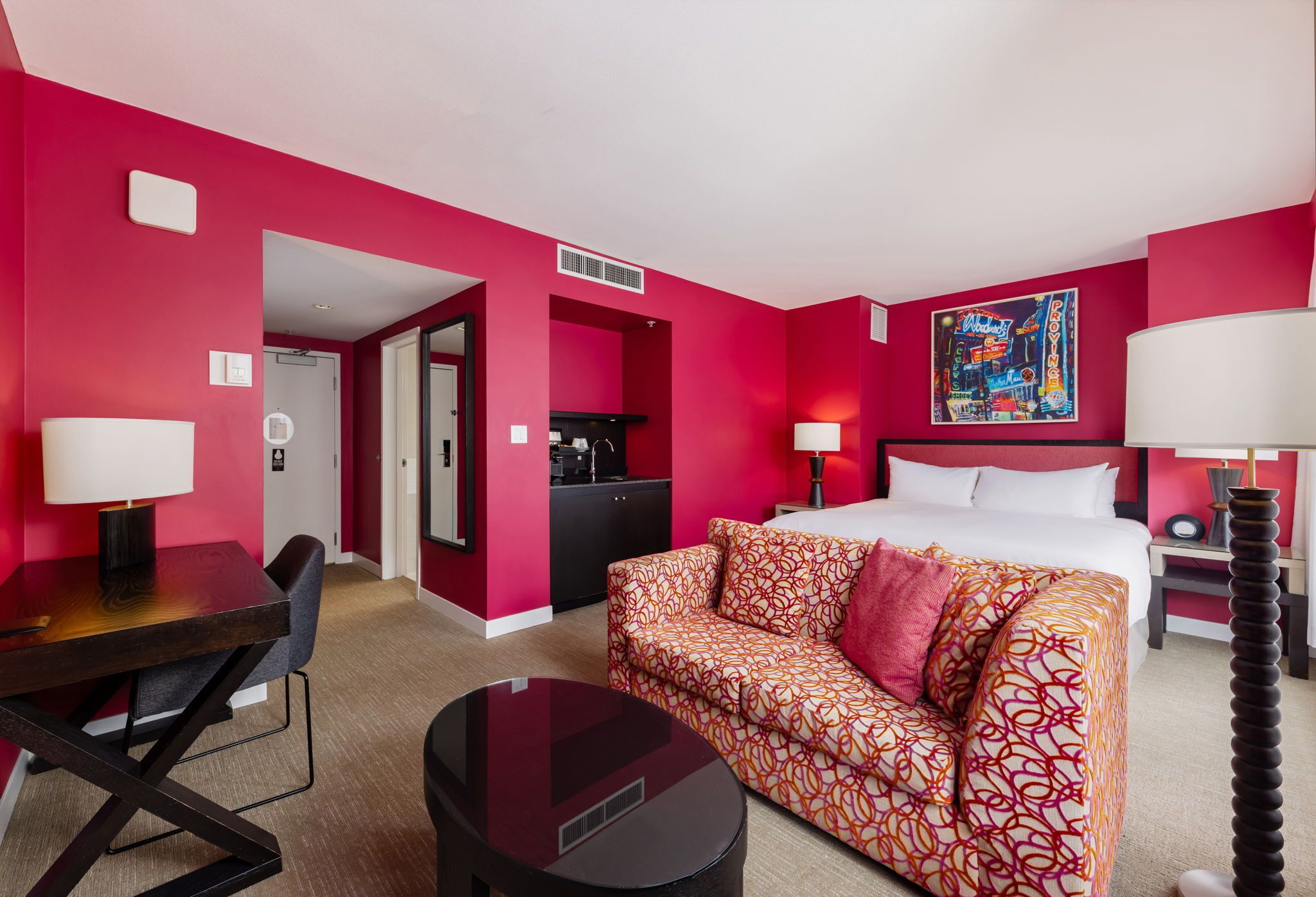 A hotel room with a bed, a sofa, a desk, and a dining table. There is a painting on the wall and a lamp on the side table.