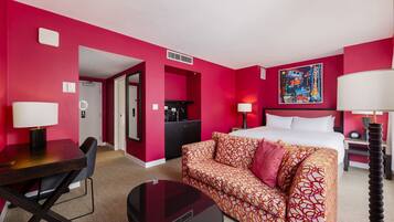 A hotel room with a bed, a sofa, a desk, and a dining table. There is a painting on the wall and a lamp on the side table.