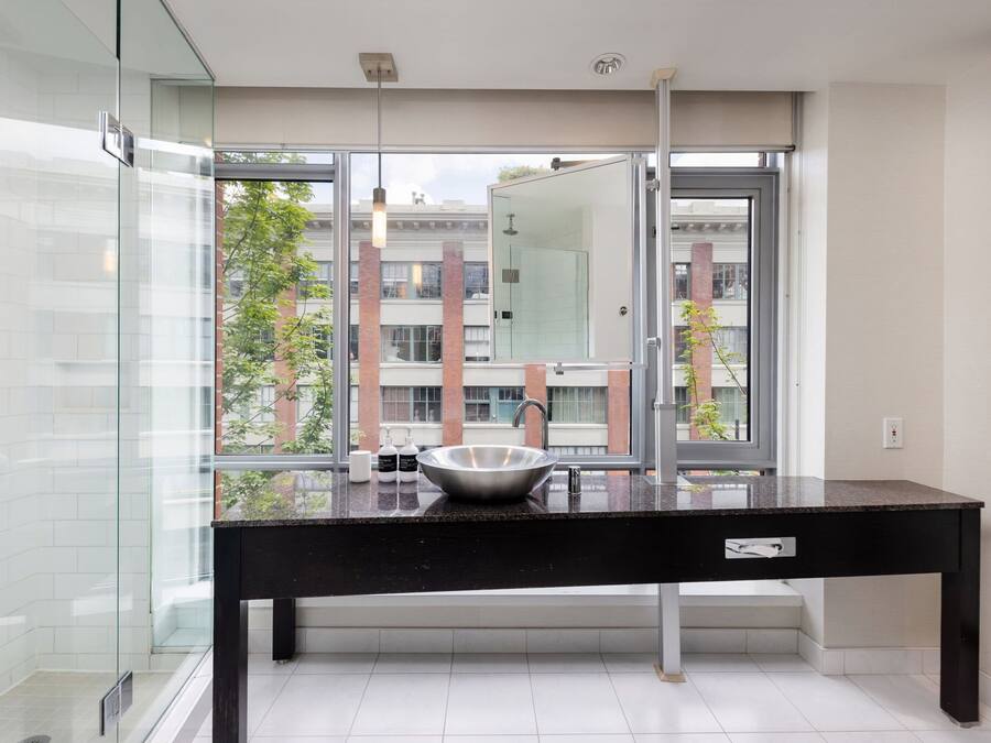 Un baño moderno con un amplio box de ducha de vidrio, encimera negra y un gran ventanal con vistas a edificios y árboles.