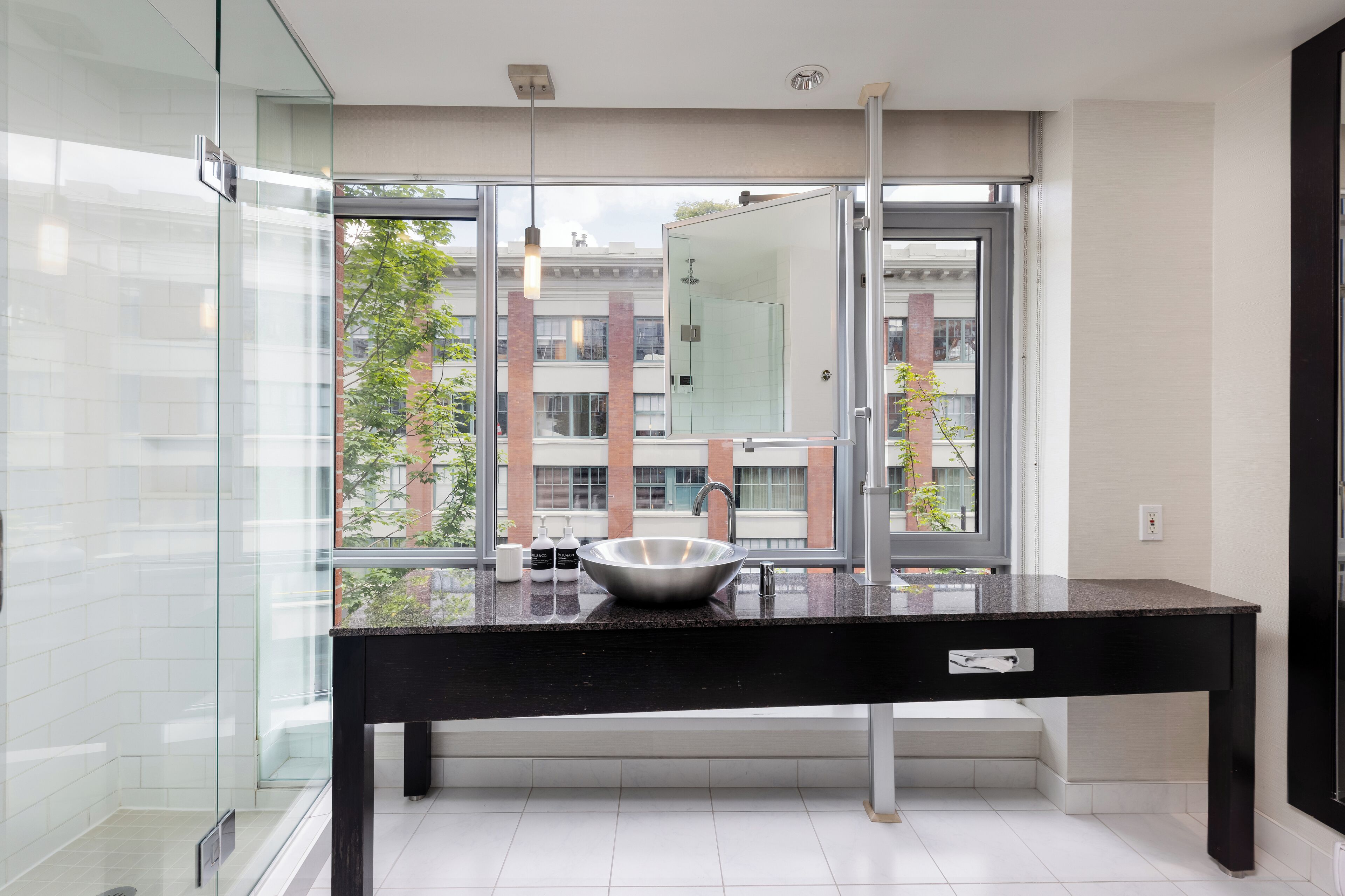 A modern bathroom with a large glass shower, a black countertop, and a view of buildings through large windows.
