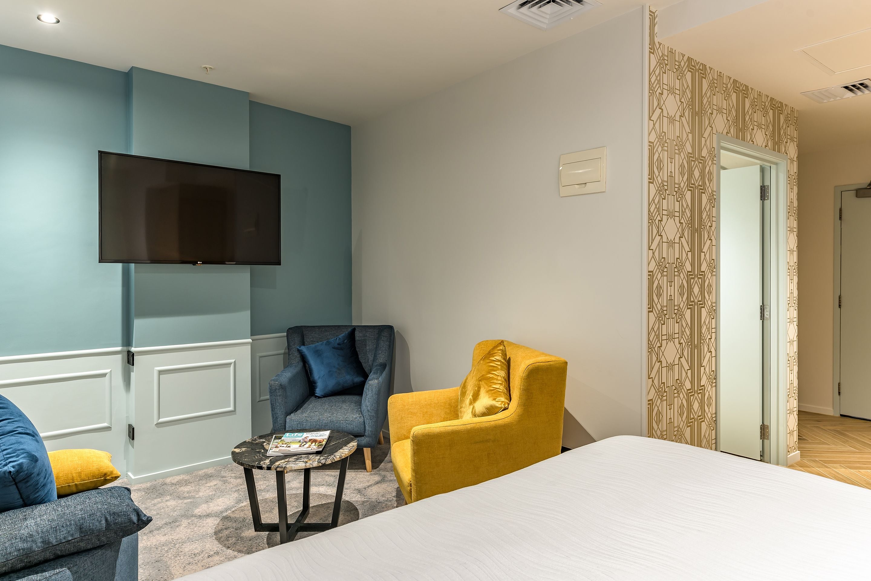 A hotel room with a large bed, a flat-screen TV mounted on the wall, two armchairs, a small table, and a patterned door.