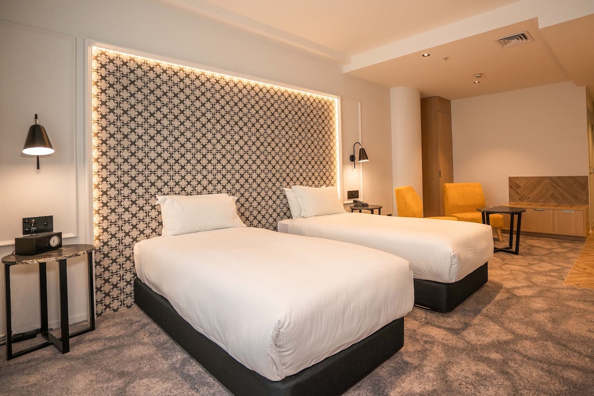 A hotel room with two beds, a small table, a chair, and a patterned headboard.