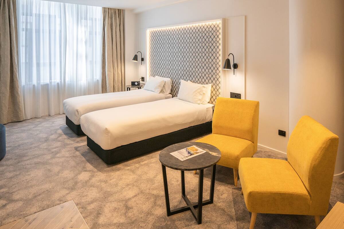 A hotel room with two beds, a small round table, a yellow armchair, and a patterned headboard.