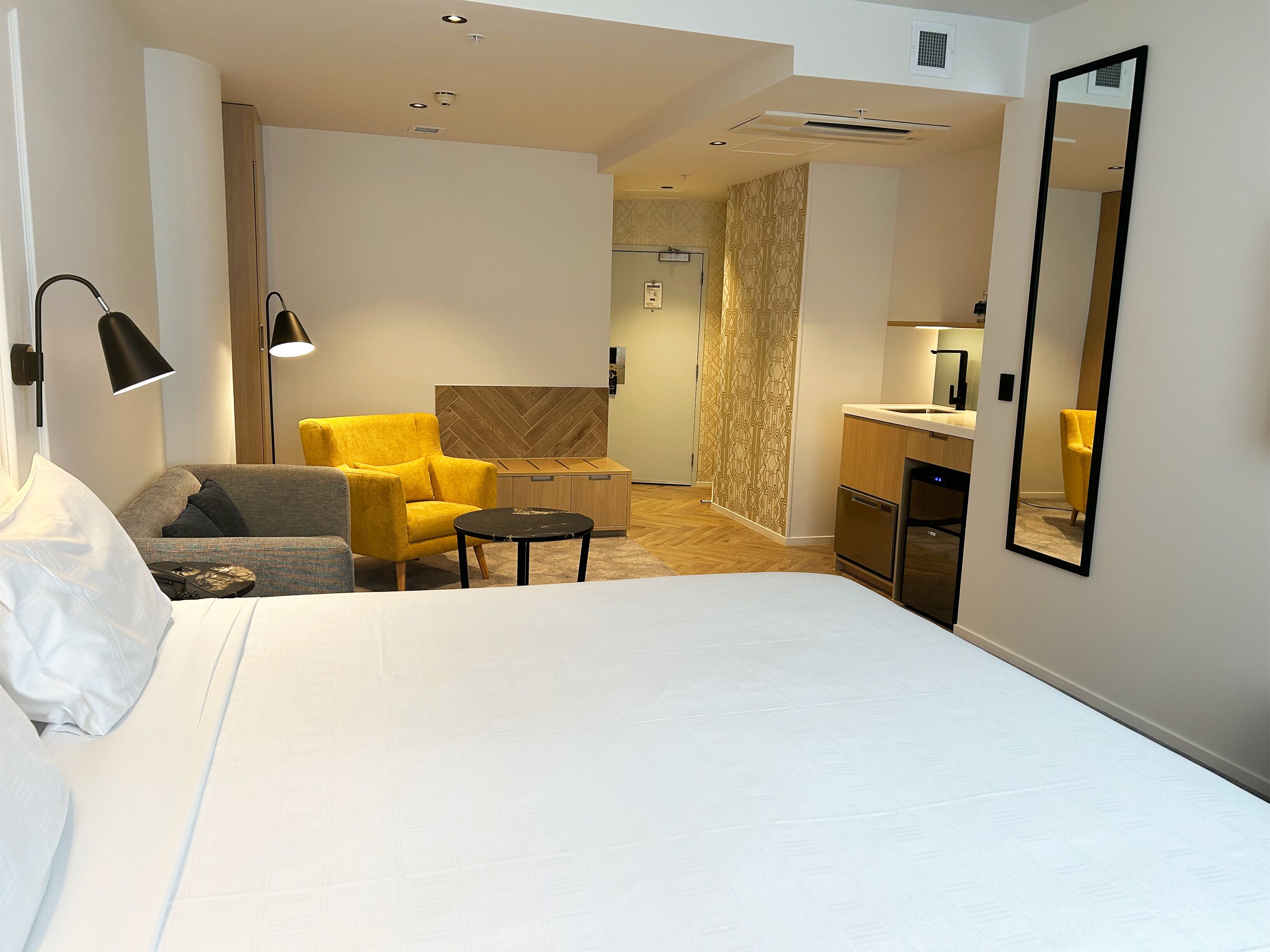 A hotel room with a bed, a sofa, a yellow armchair, a small table, a mirror, and a kitchenette.