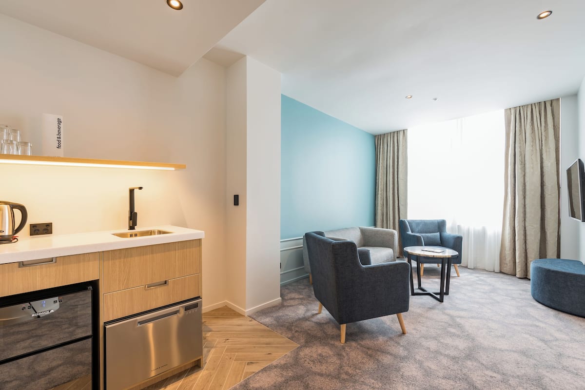 A modern hotel room with a kitchenette, a seating area with a sofa and armchairs, and a large window with curtains.