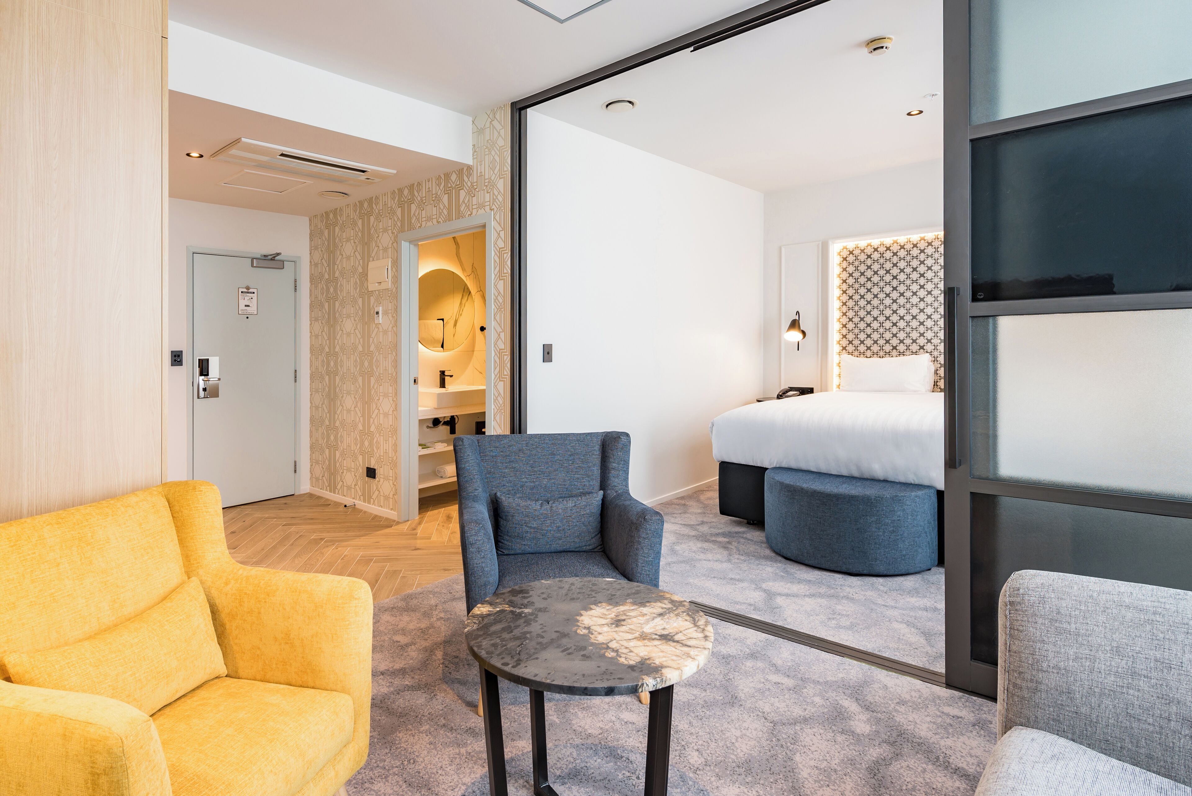 Ein Hotelzimmer mit einer Couch, einem Sessel und einem runden Tisch. Ein Bett mit gemustertem Kopfteil und ein kleiner Hocker sind ebenfalls vorhanden. Eine Tür führt zu einem Badezimmer mit Spiegel und Handtuchhalter.