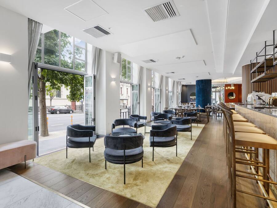 A modern hotel lobby with a long bar area, seating, and large windows offering views of the street.