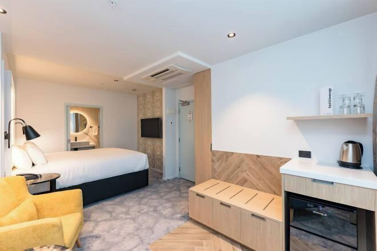 A modern hotel room with a bed, a sofa, a kitchenette, and a small dining area.